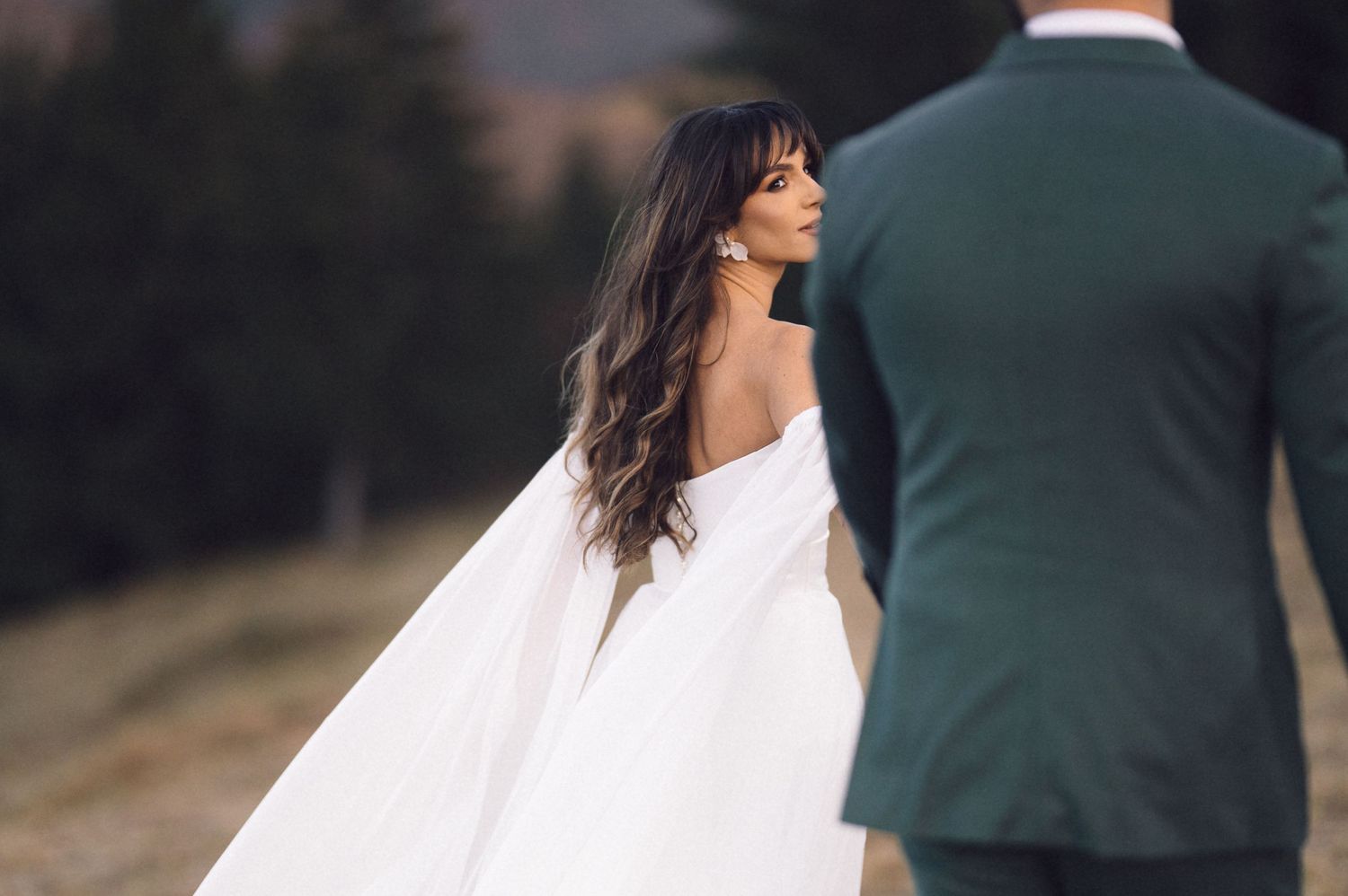 Bride in white dress walks with groom in dark suit along outdoor path during wedding photo session.