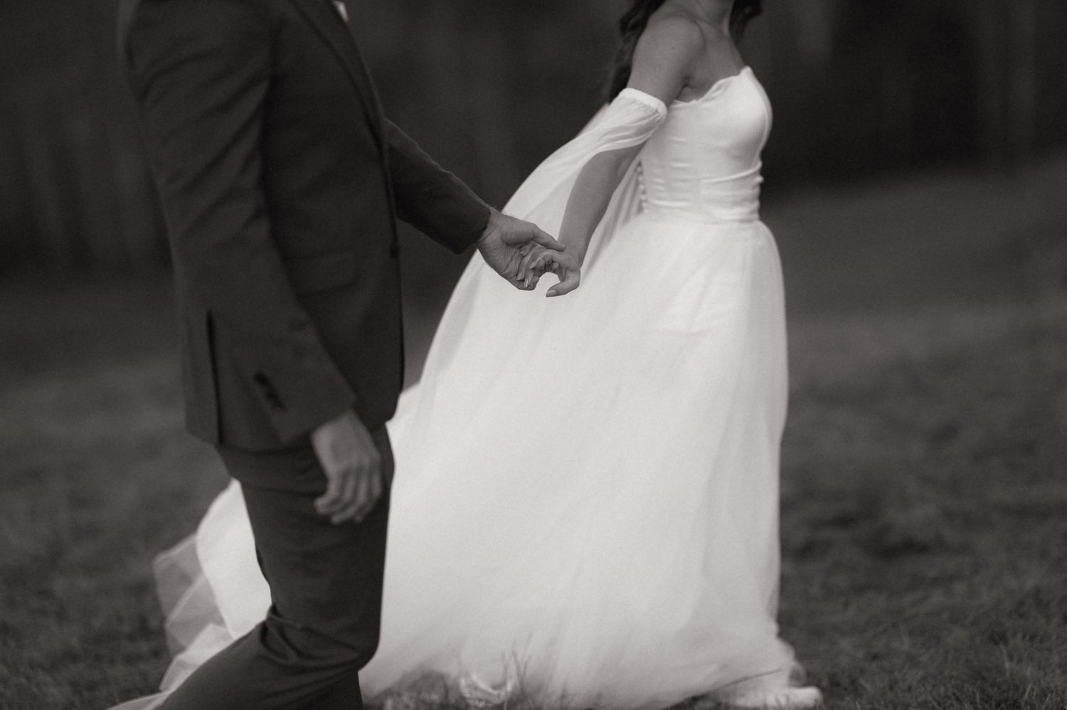 Black and white photo showing the bottom half of a couple holding hands, with bride in flowing white gown.