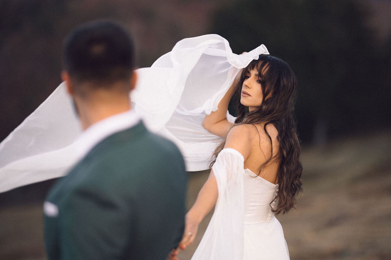 A bride in a white wedding dress with flowing veil stands outdoors while her veil catches the wind.
