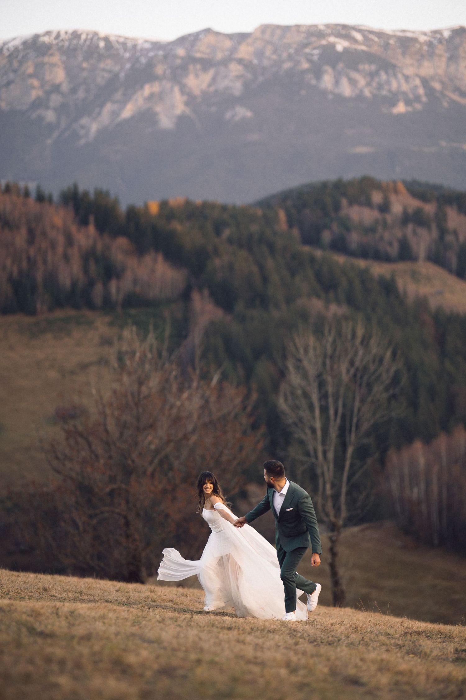 A couple walks along a scenic mountain ridge with dramatic peaks and autumn foliage in the background.
