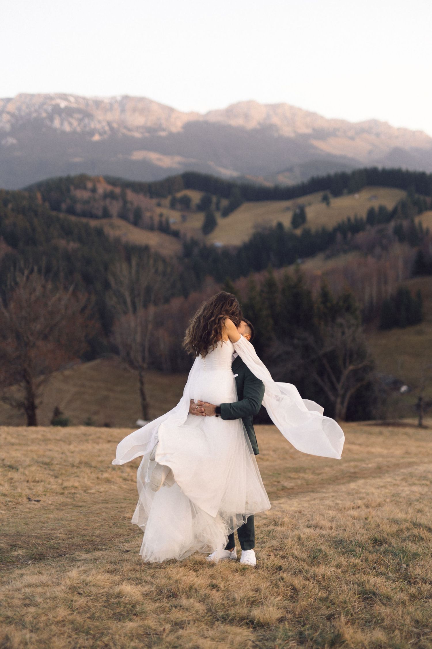 A couple in wedding attire dances together on a scenic mountain meadow with snow-capped peaks in the distance.