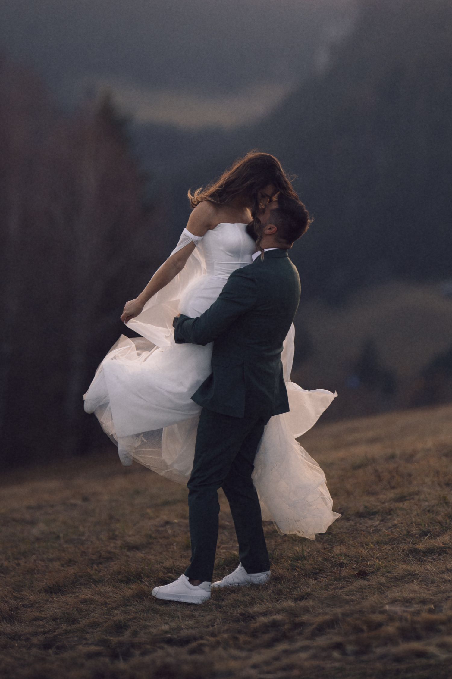 A couple in wedding attire embraces and dances together on a hillside at dusk creating a romantic silhouette.