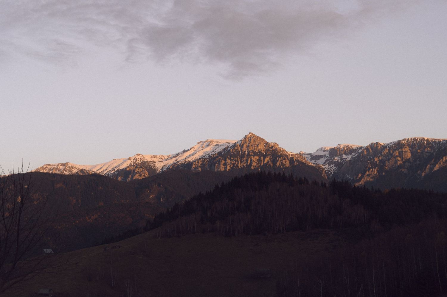 Panoramic mountain landscape at sunset with snow-capped peaks glowing in warm golden light against darkening sky.