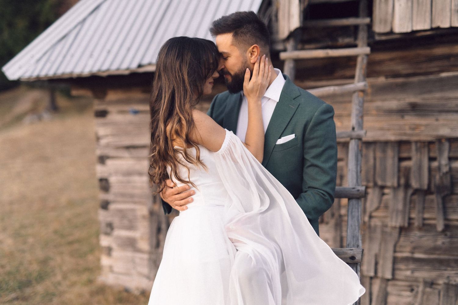 A romantic couple in wedding attire embrace in front of rustic wooden buildings during an outdoor photoshoot.