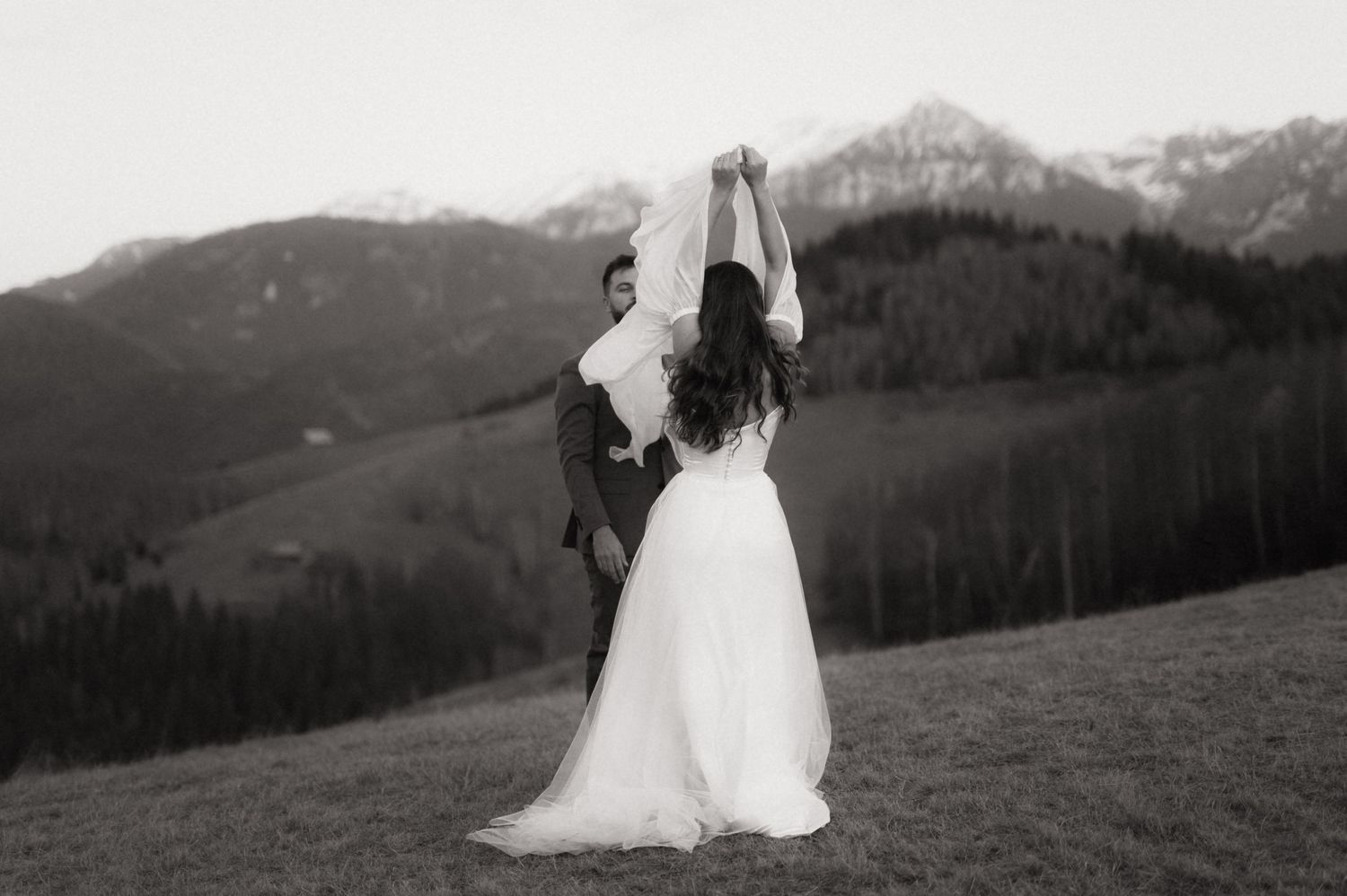 A couple in wedding attire embraces on a scenic mountain ridge during sunset with dramatic landscapes in the background.