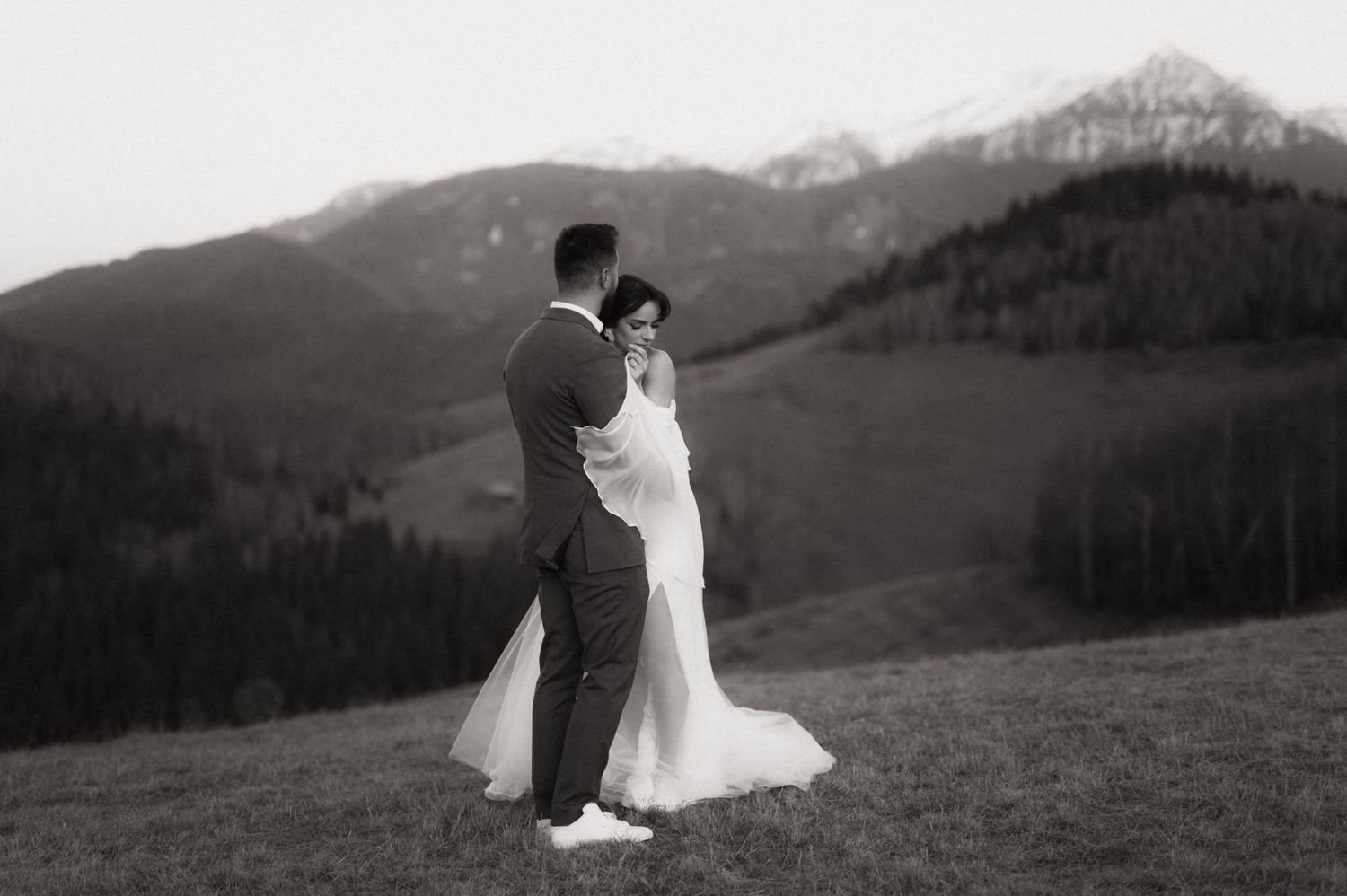 A couple in wedding attire walks across a scenic mountain meadow with dramatic mountain peaks in the background.