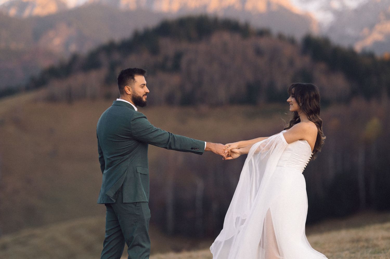 A couple in wedding attire poses together in front of a scenic mountain landscape during sunset.