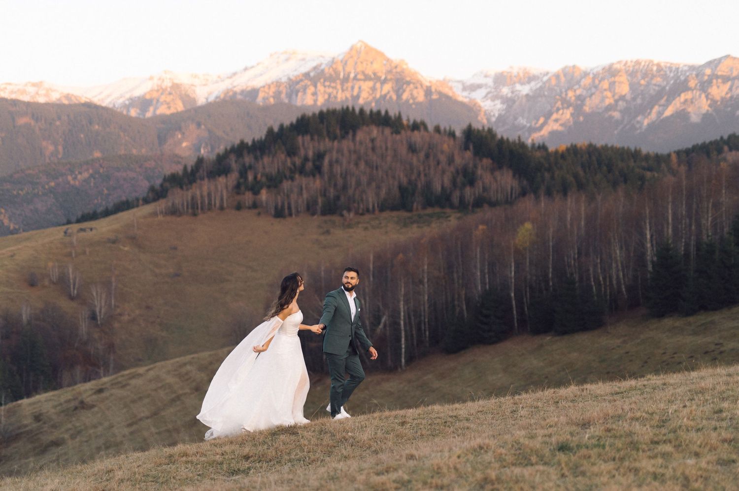 A couple in wedding attire stands together against a scenic mountain landscape at sunset.