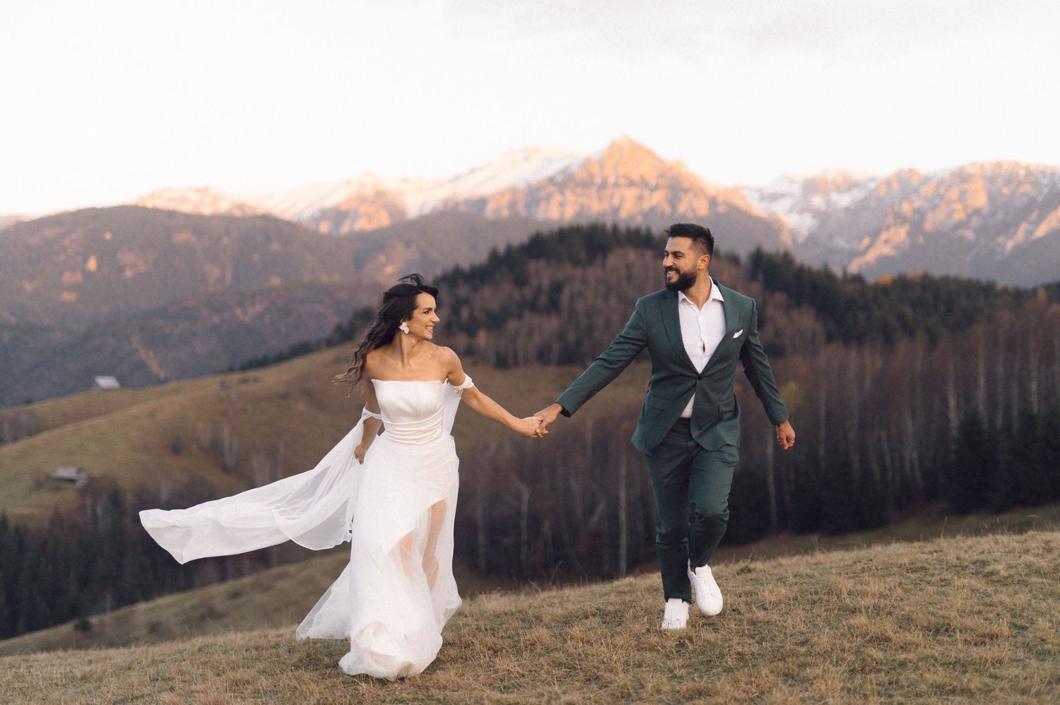 Couple in wedding attire poses against stunning mountain landscape at sunset with flowing white dress in the breeze.
