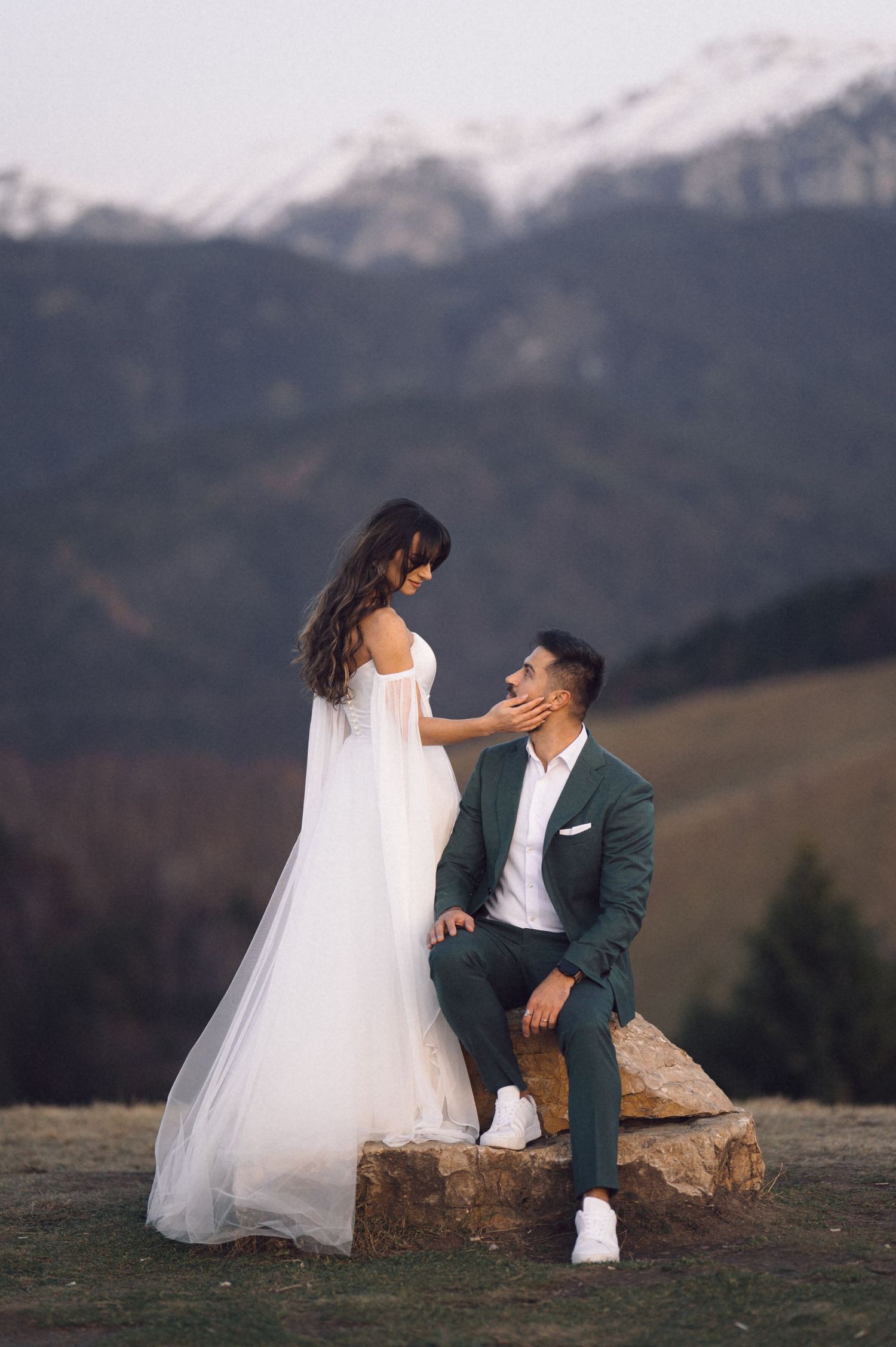 A romantic wedding photo in the mountains with a couple posing on a rock, snow-capped peaks in the background.