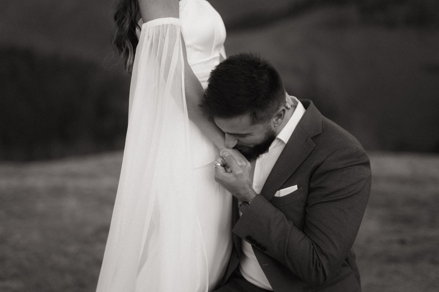 A tender black and white wedding moment captured outdoors with a flowing white veil and elegant formal attire.