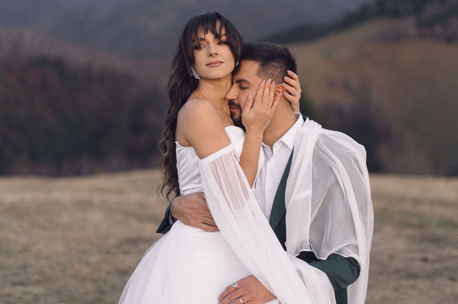 Romantic couple in white wedding attire embrace on mountainside during sunset photoshoot.