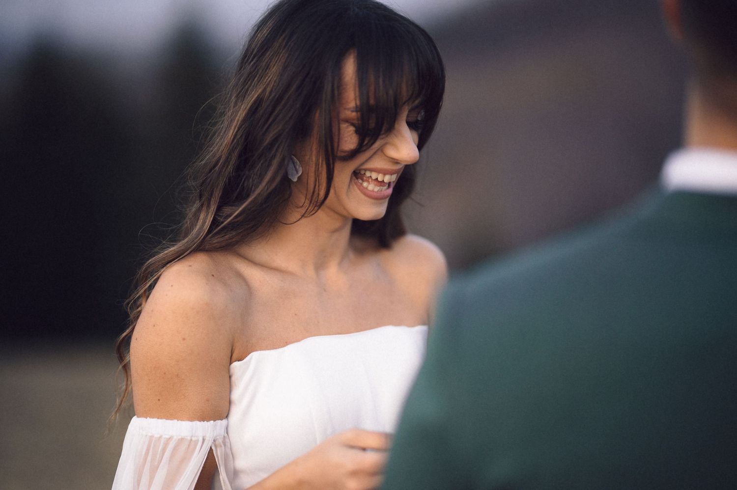 A bride in a white off-shoulder wedding dress smiles joyfully while walking outdoors.