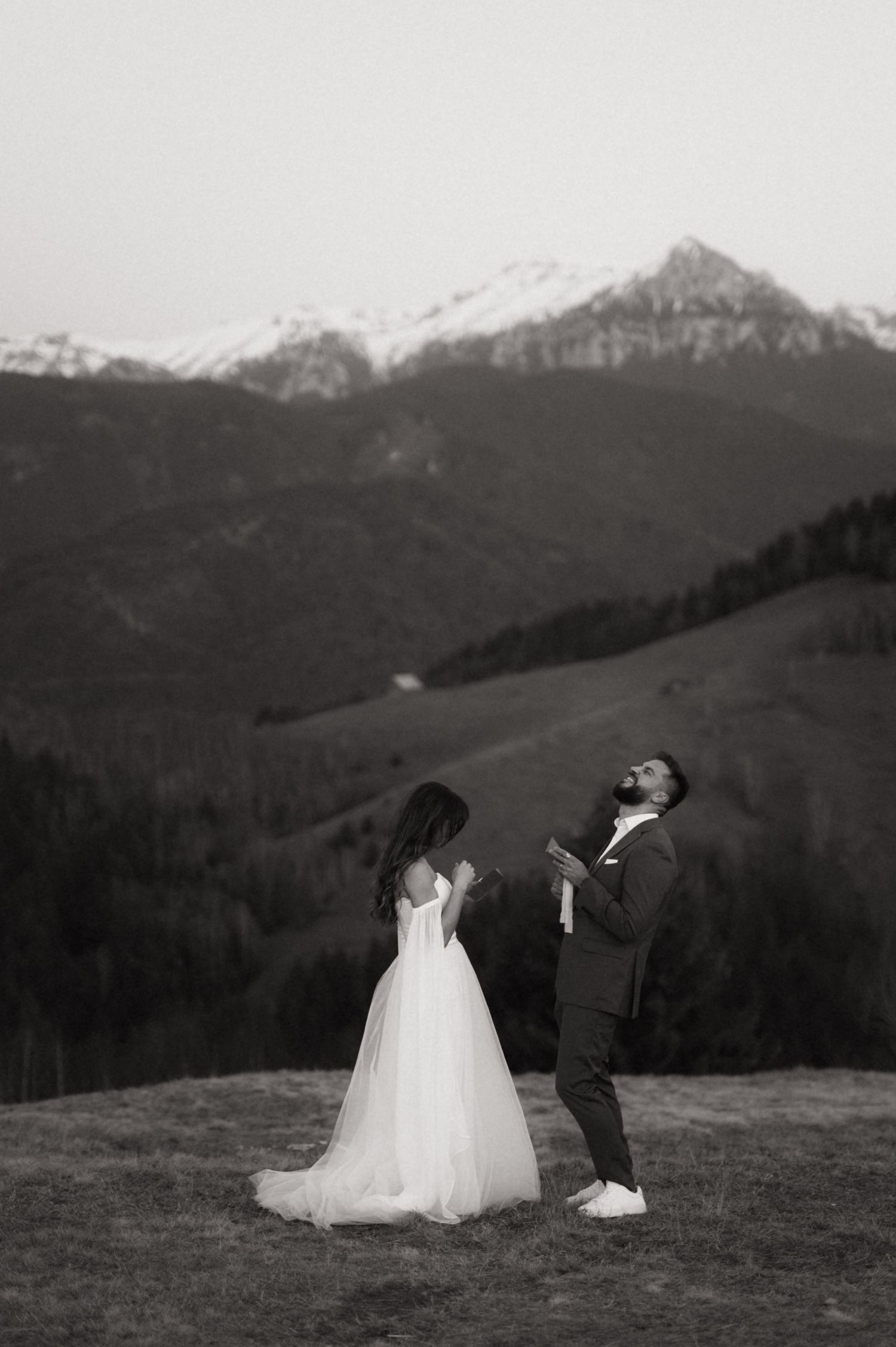 A bride and groom pose playfully against a backdrop of snow-capped mountains in a black and white wedding photo.