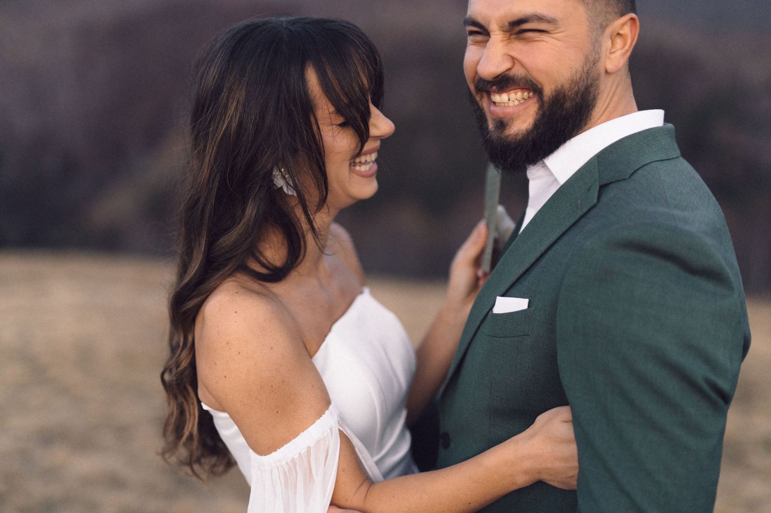 A couple in wedding attire embraces and smiles while standing outdoors in a natural setting.