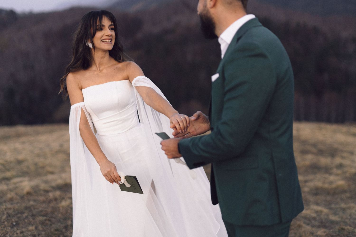 A couple in wedding attire share a joyful moment together on a hilltop during their outdoor mountain ceremony.