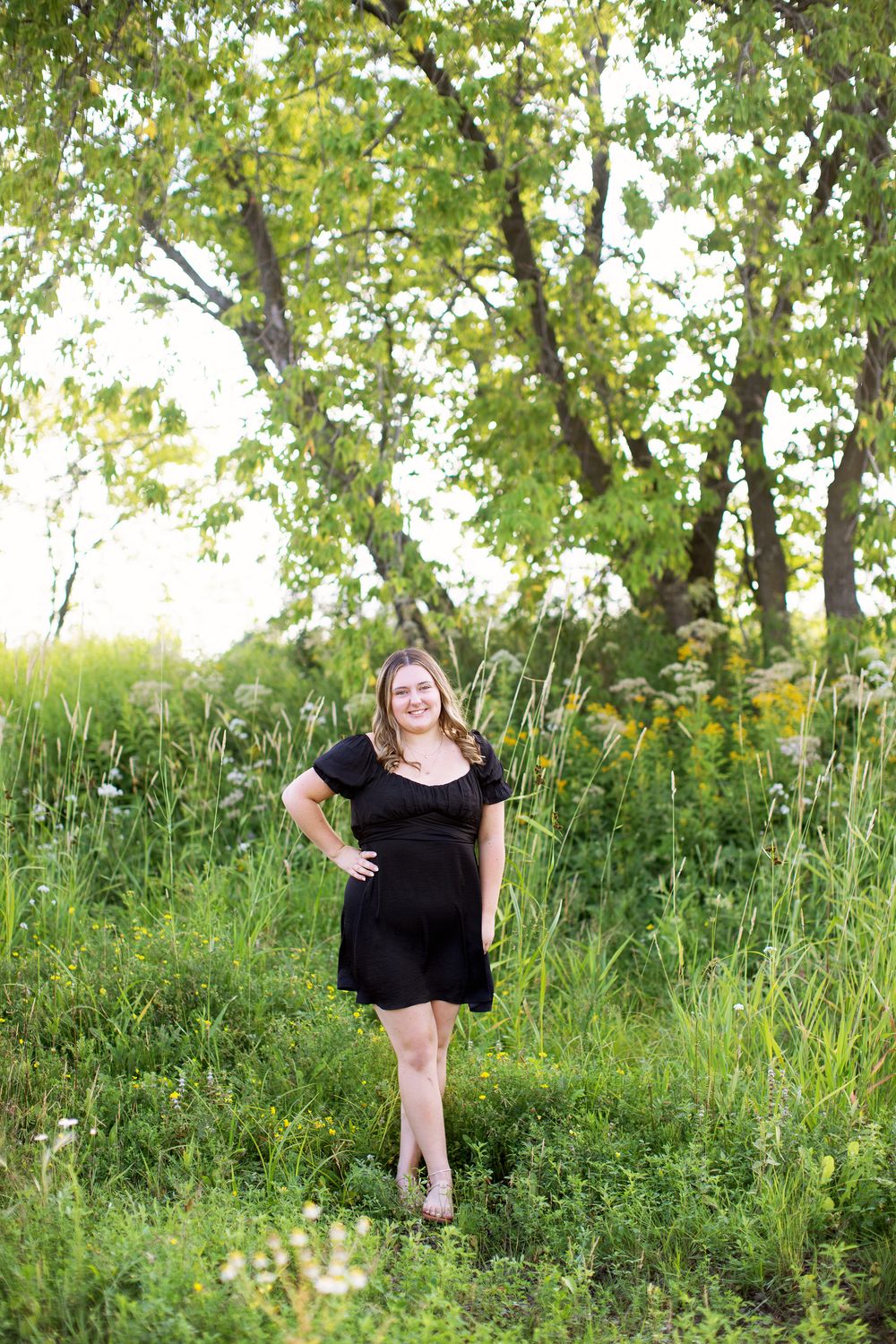 A person in a black dress poses in a lush green field with trees in the background on a sunny day.