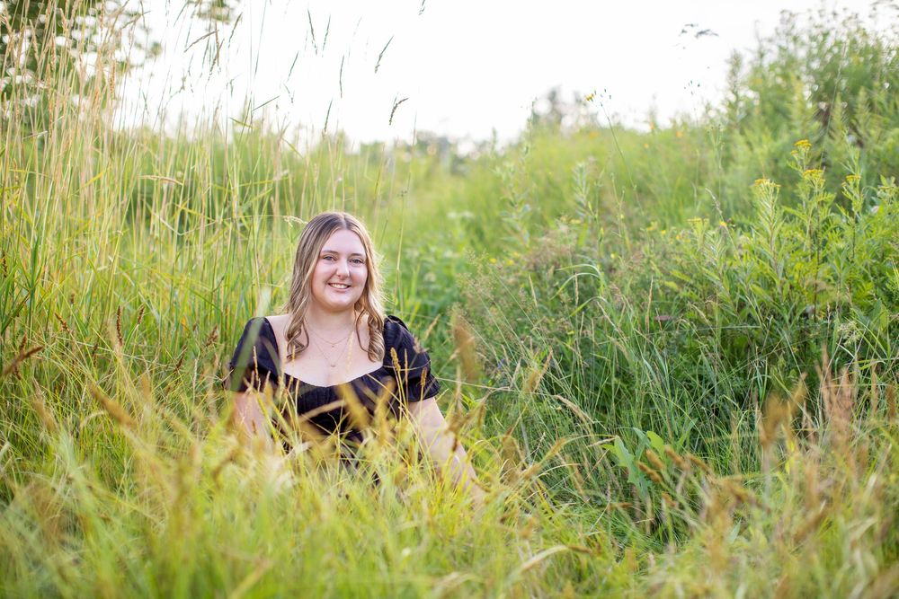 Someone wearing a black dress sits among tall green grass and wildflowers in a sunny meadow.