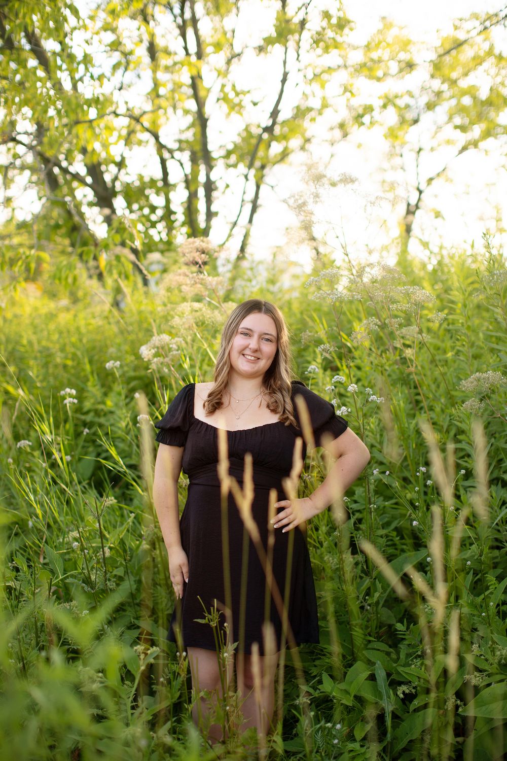 A person in a black dress stands in a sunny meadow filled with tall wildflowers and grasses.