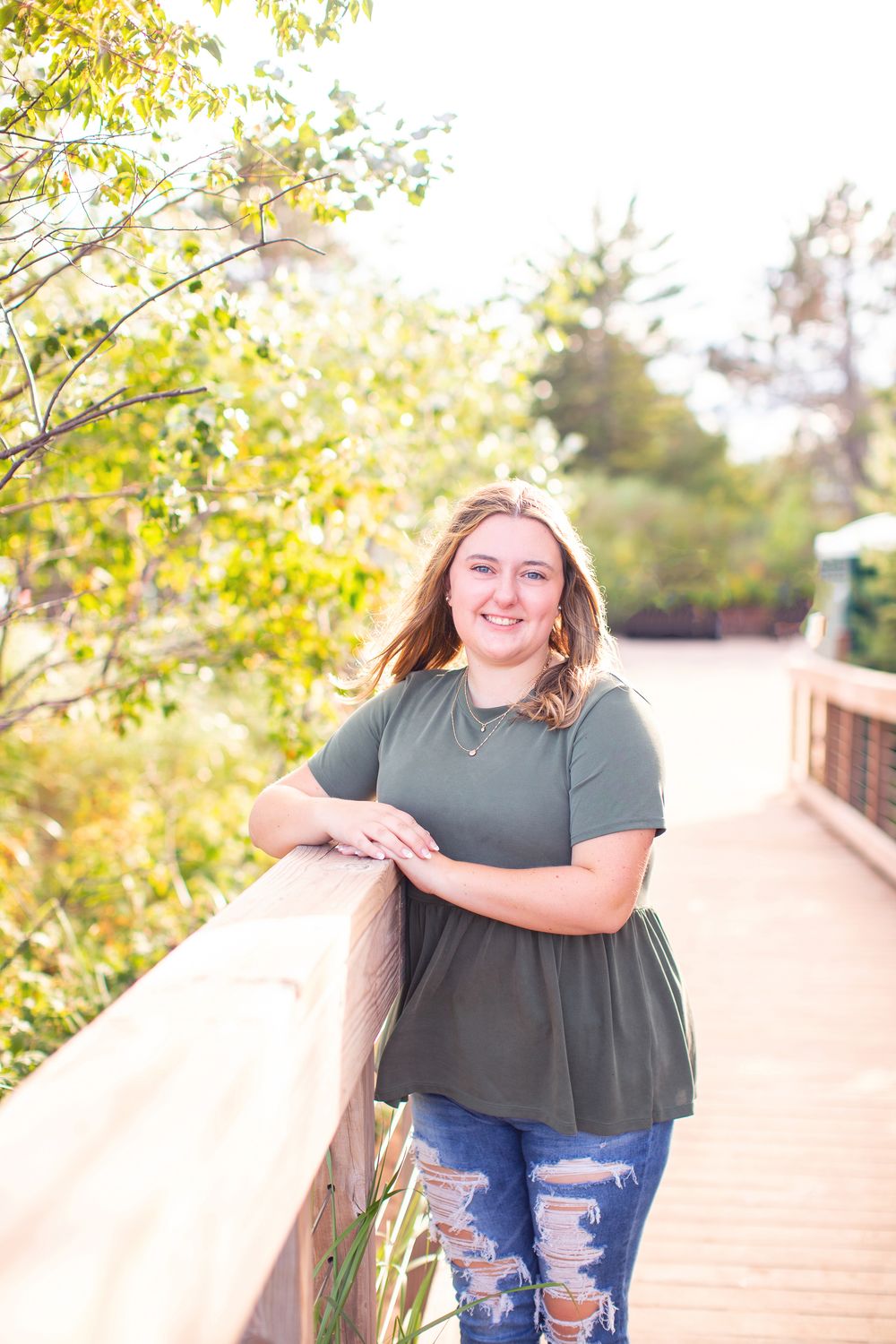 A person in a green top and ripped jeans poses outdoors on a wooden deck surrounded by autumn foliage.