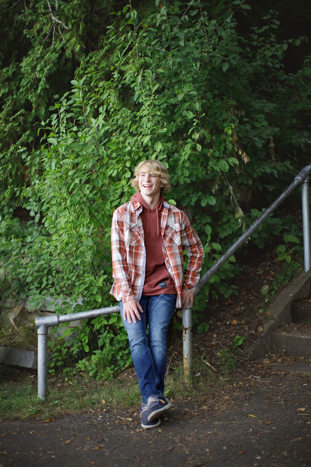 A person in a plaid shirt and jeans sits casually on a metal railing along a wooded path.