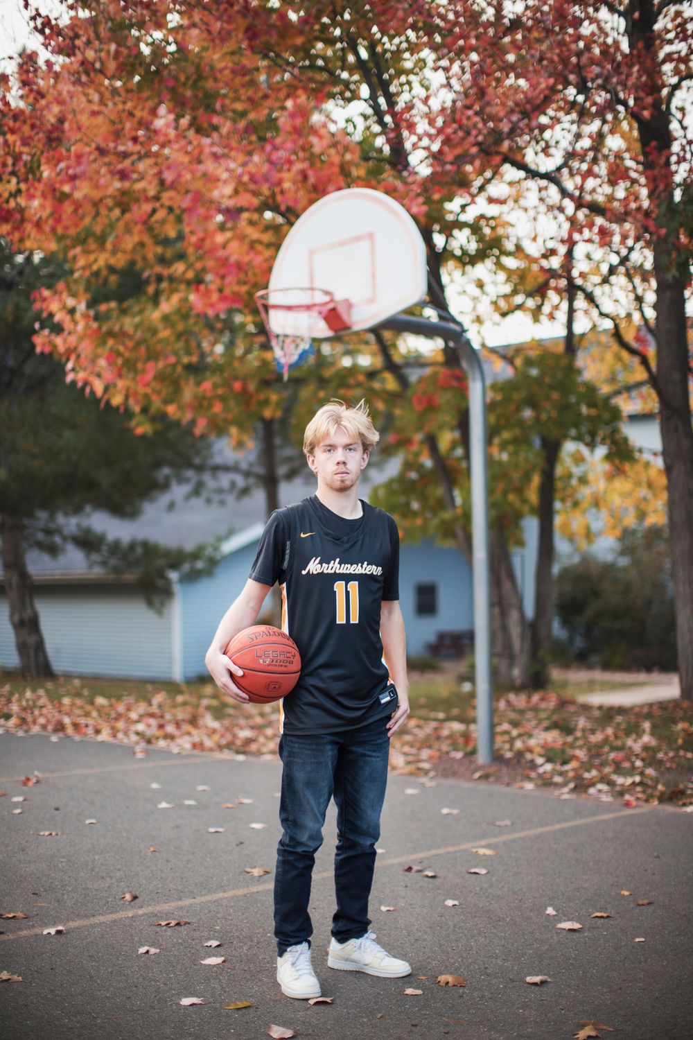 A basketball player dribbles on an outdoor court surrounded by autumn trees with colorful red foliage.