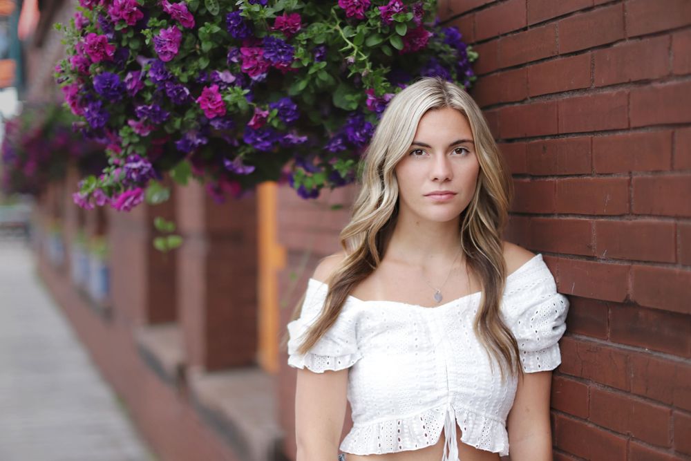 A person in a white crop top stands against a brick wall with purple flowers blooming overhead.