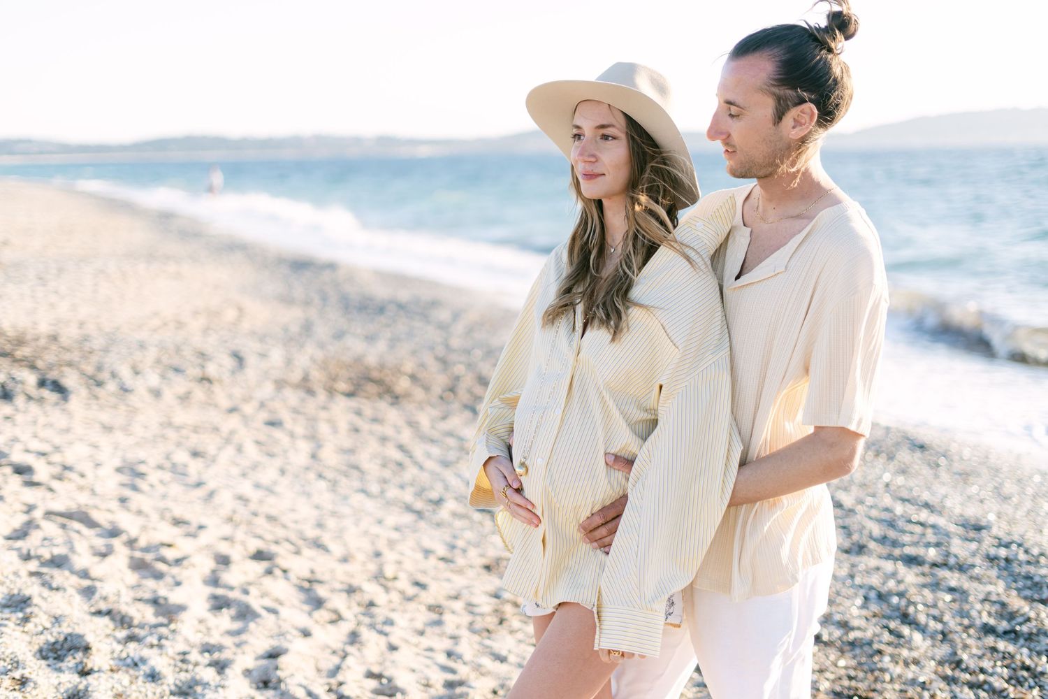 A couple in light colored clothes embraces while standing on a sunny beach with ocean waves in the background.