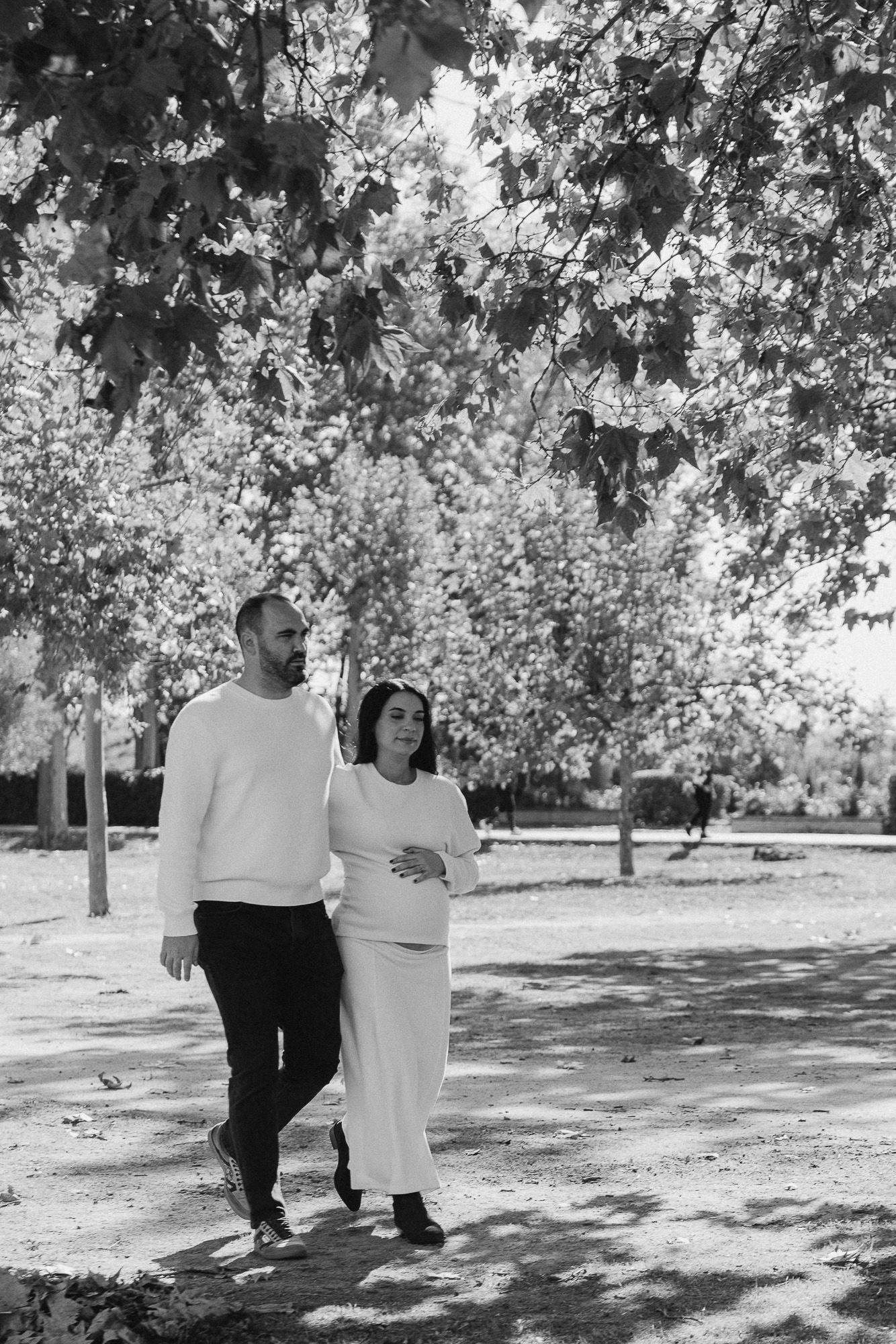 A couple walking together in a park on a sunny day, with trees and dappled light creating a romantic black and white scene.
