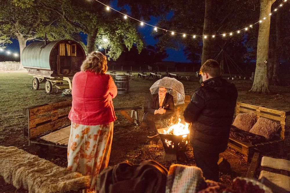 People gather around a cozy campfire and string lights at night in an outdoor seating area with rustic log benches.