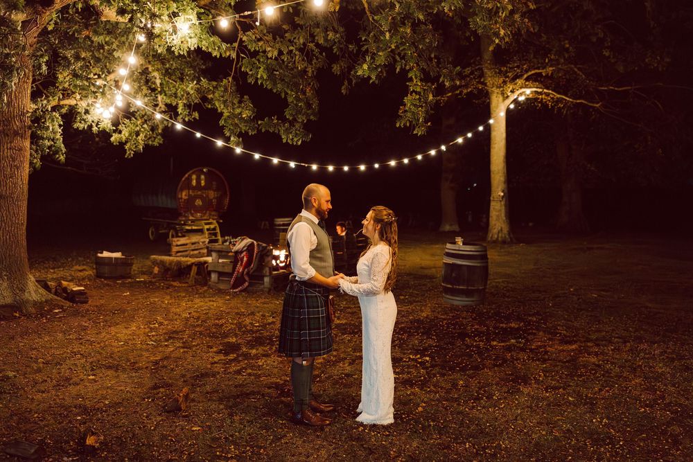 String lights illuminate a romantic outdoor evening wedding dance under tall trees in a forest setting.