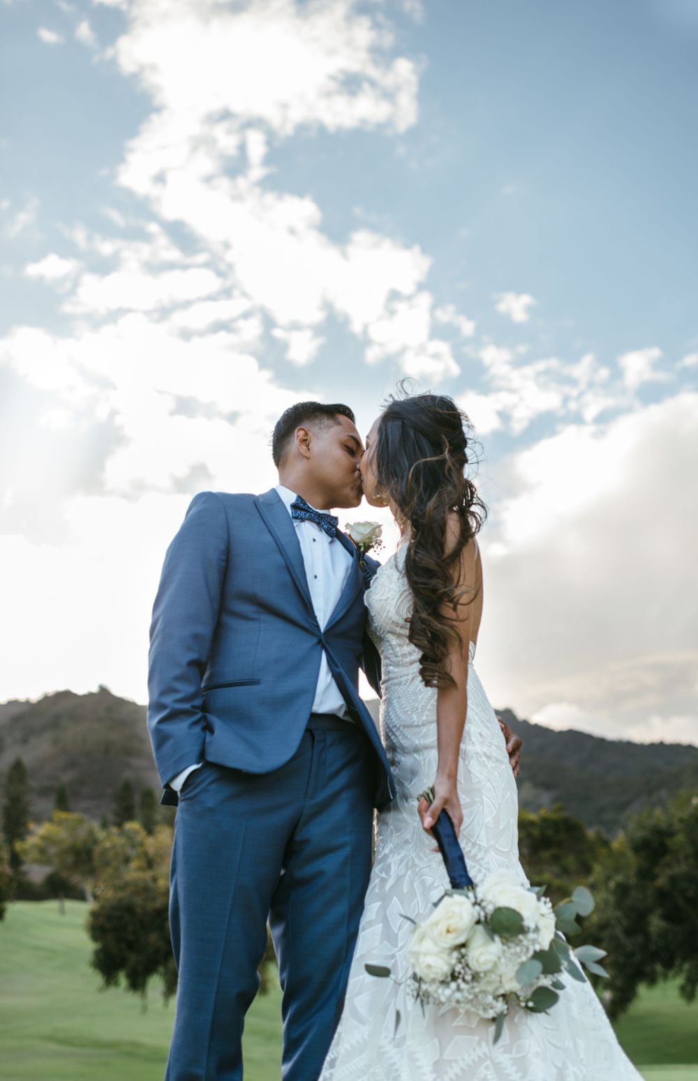 A couple in wedding attire embraces against a cloudy mountain backdrop.