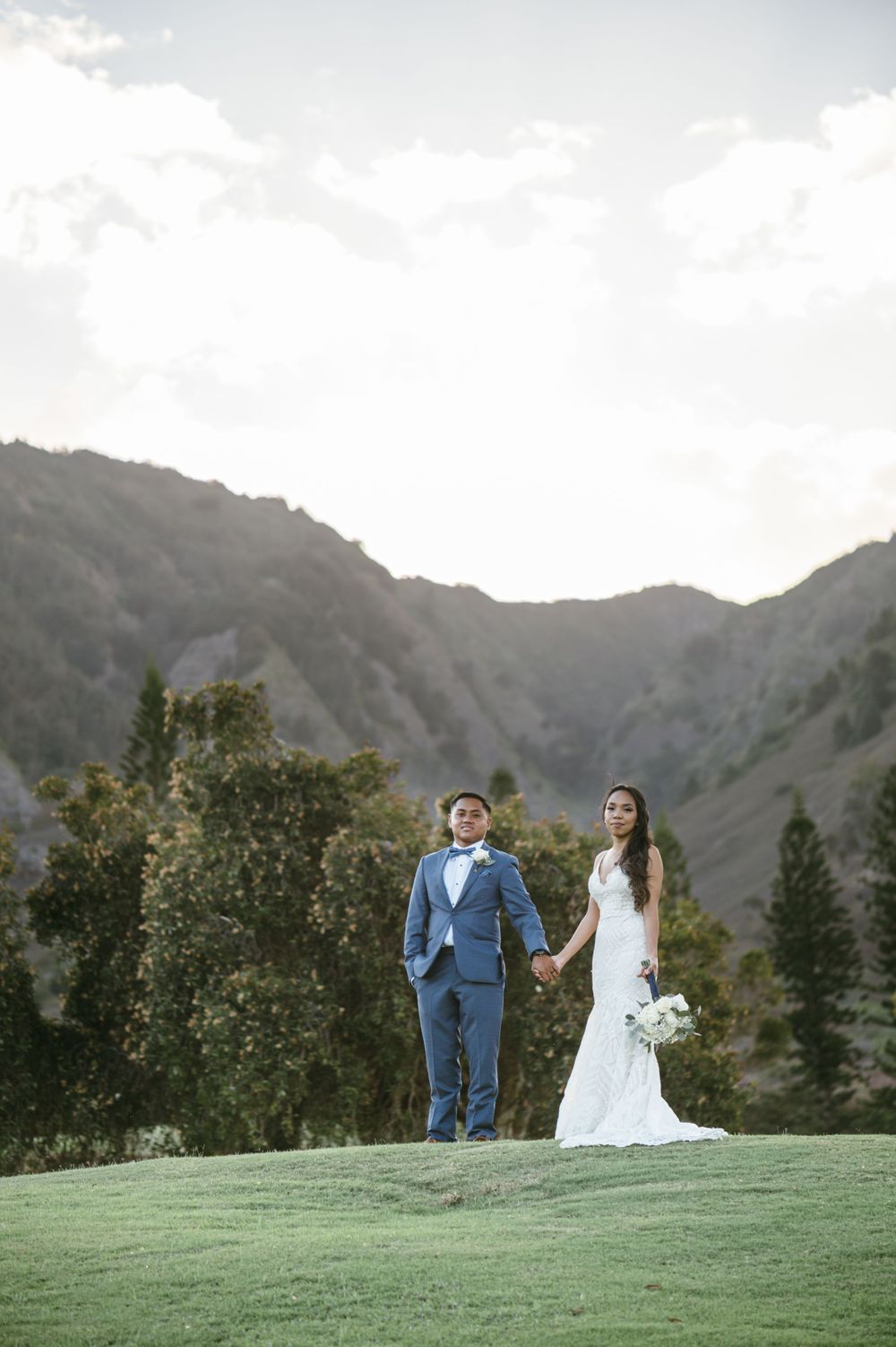 A couple in formal attire stands together on a scenic mountain overlook.