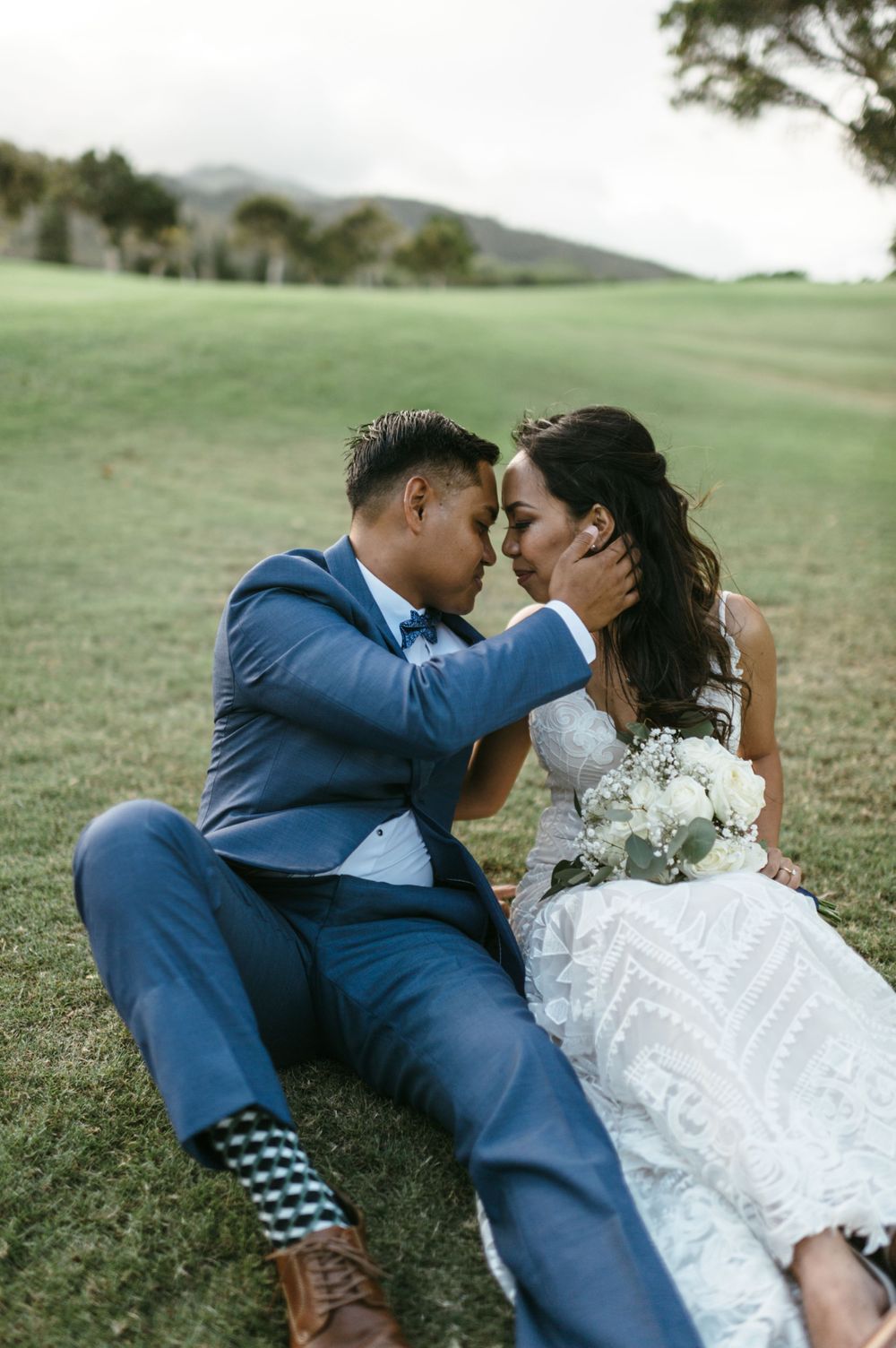 A couple in wedding attire shares an intimate moment lying on green grass.