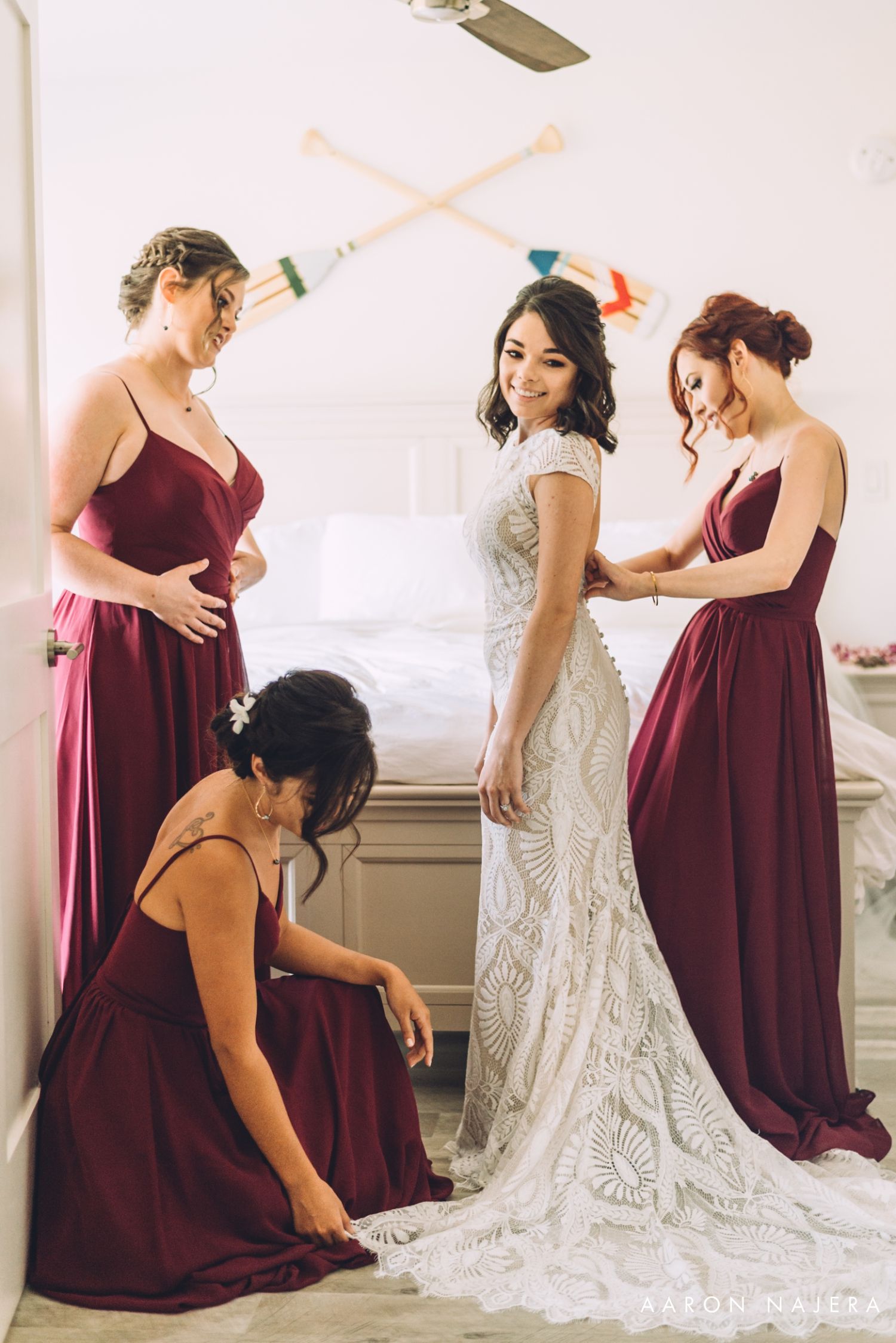 Bridesmaids in burgundy dresses help the bride with her lace wedding gown during preparation.