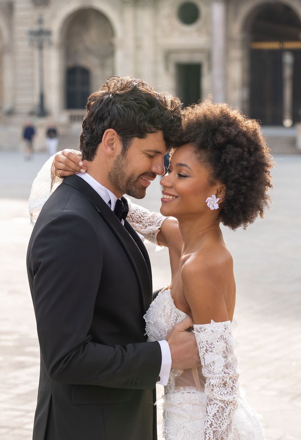 A romantic wedding couple embraces in an elegant outdoor setting with classical architecture in the background.