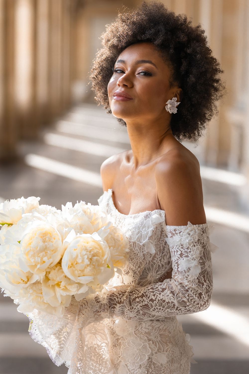 A bride in an off-shoulder lace wedding dress poses elegantly in natural window light with cream-colored flowers.