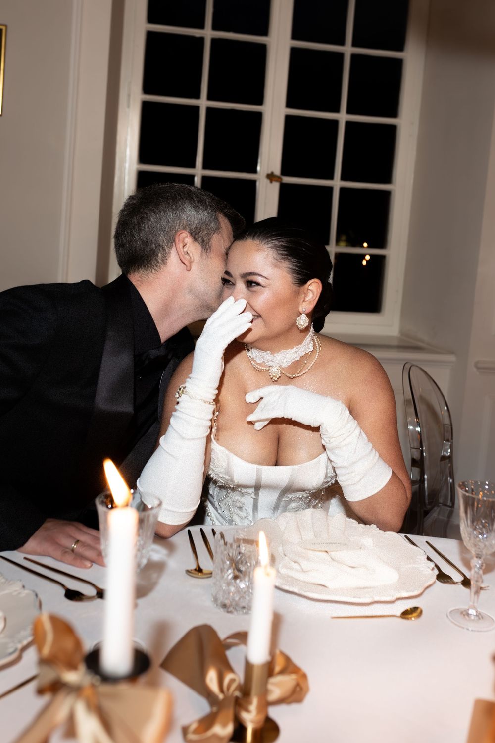 A couple shares an intimate moment during a candlelit dinner reception with white tablecloths and elegant place settings.