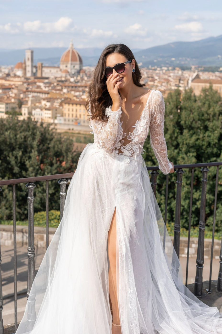 A bride in a flowing white lace gown poses at a scenic overlook with Florence's iconic Duomo cathedral visible in the distance.