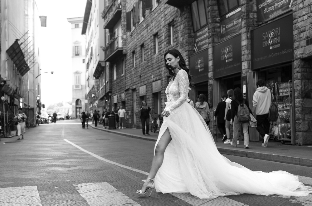 A bride in a flowing white wedding gown poses on a cobblestone street in an urban setting.