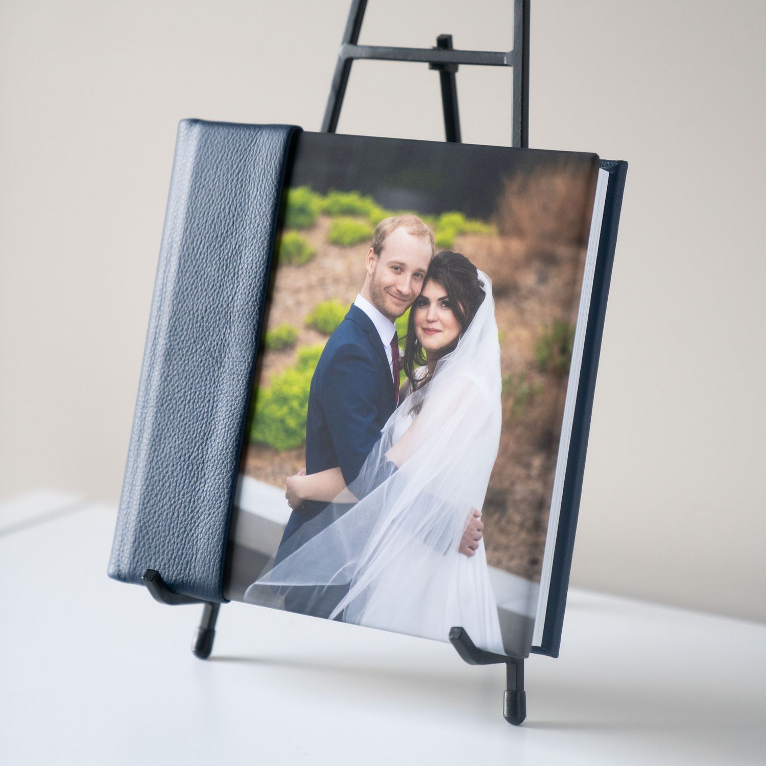 A wedding album displayed on a black easel stand shows a couple embracing outdoors in formal attire with a flowing white veil.