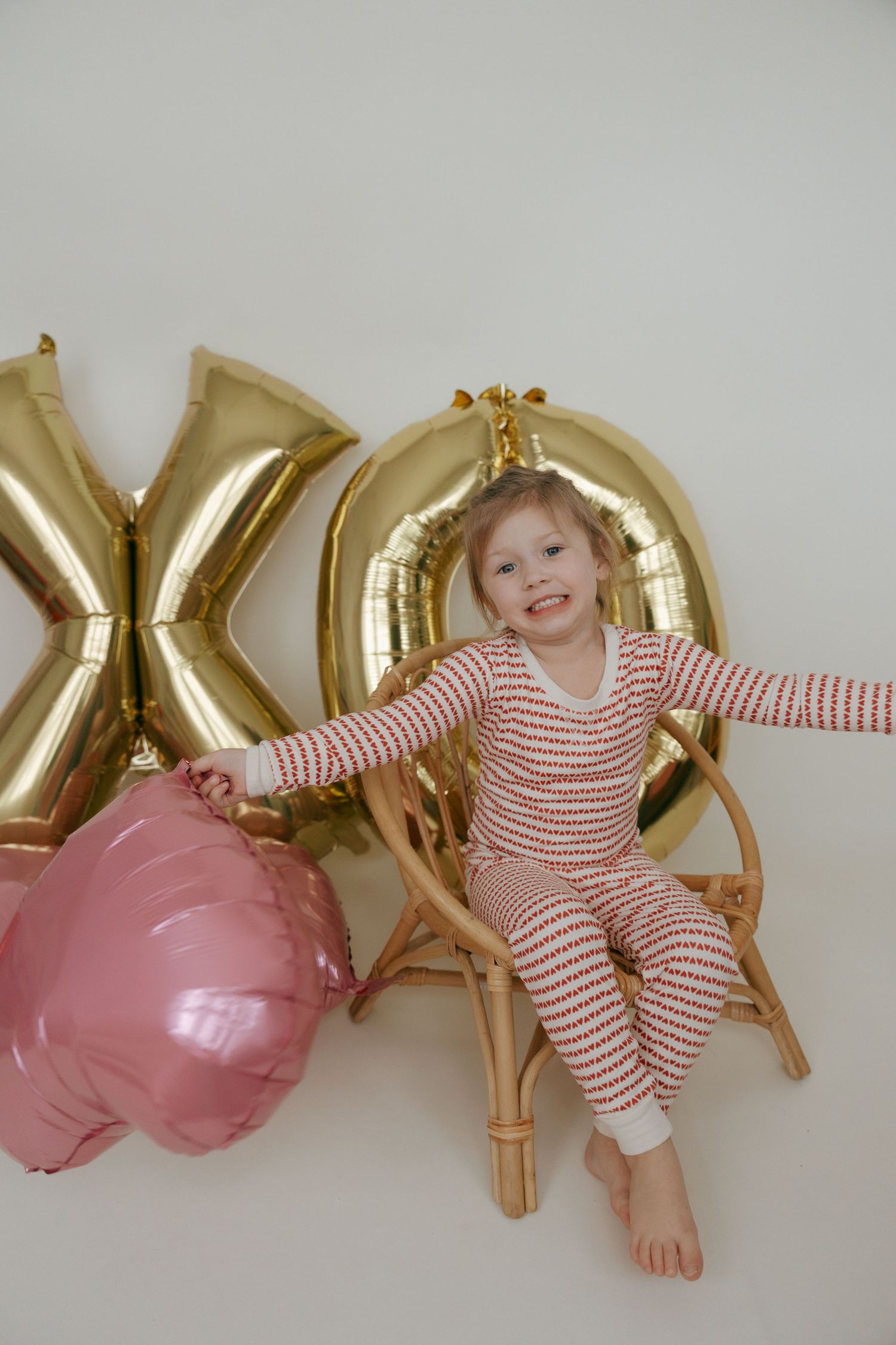 Child in red and white striped pajamas poses playfully with gold letter balloons and pink decorations against white wall.