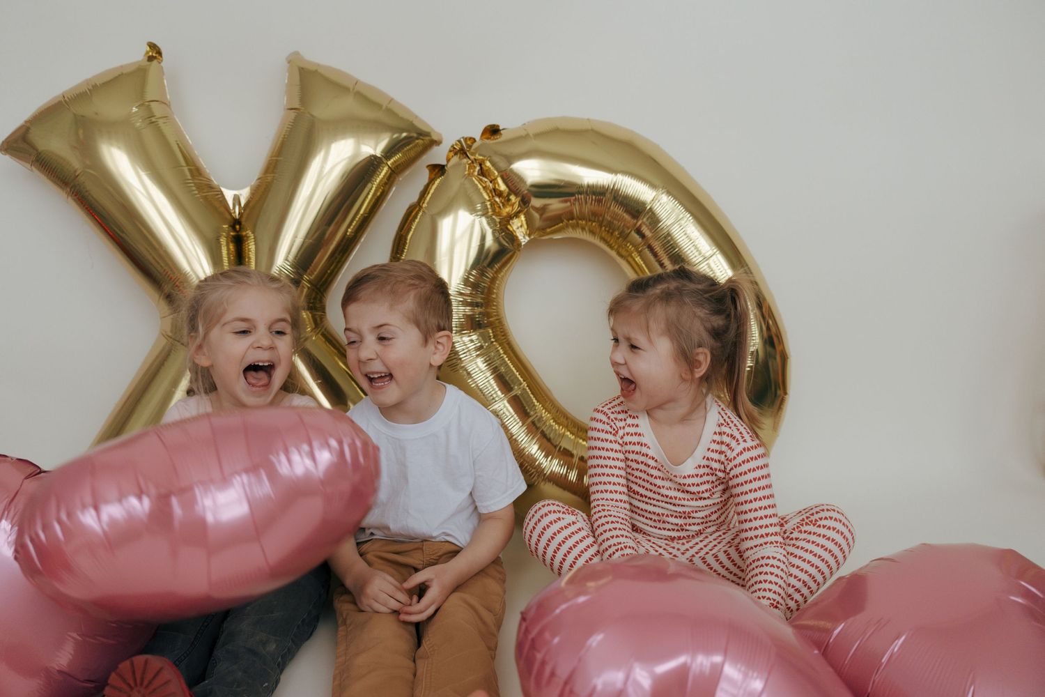 Children in striped shirts play and laugh while surrounded by pink and gold balloon decorations at a party.