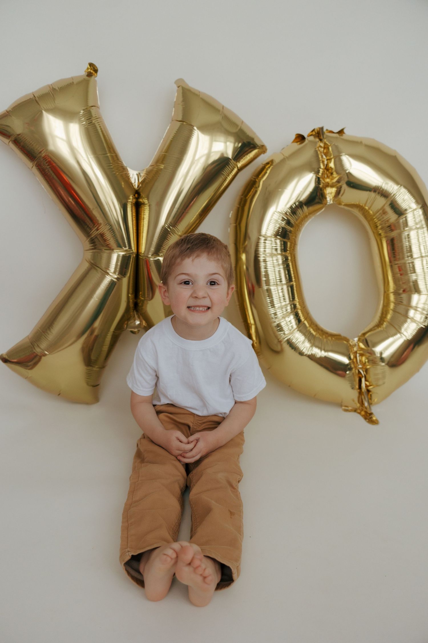A young child sits against a white wall with large gold XO letter balloons for Valentine's Day decor.