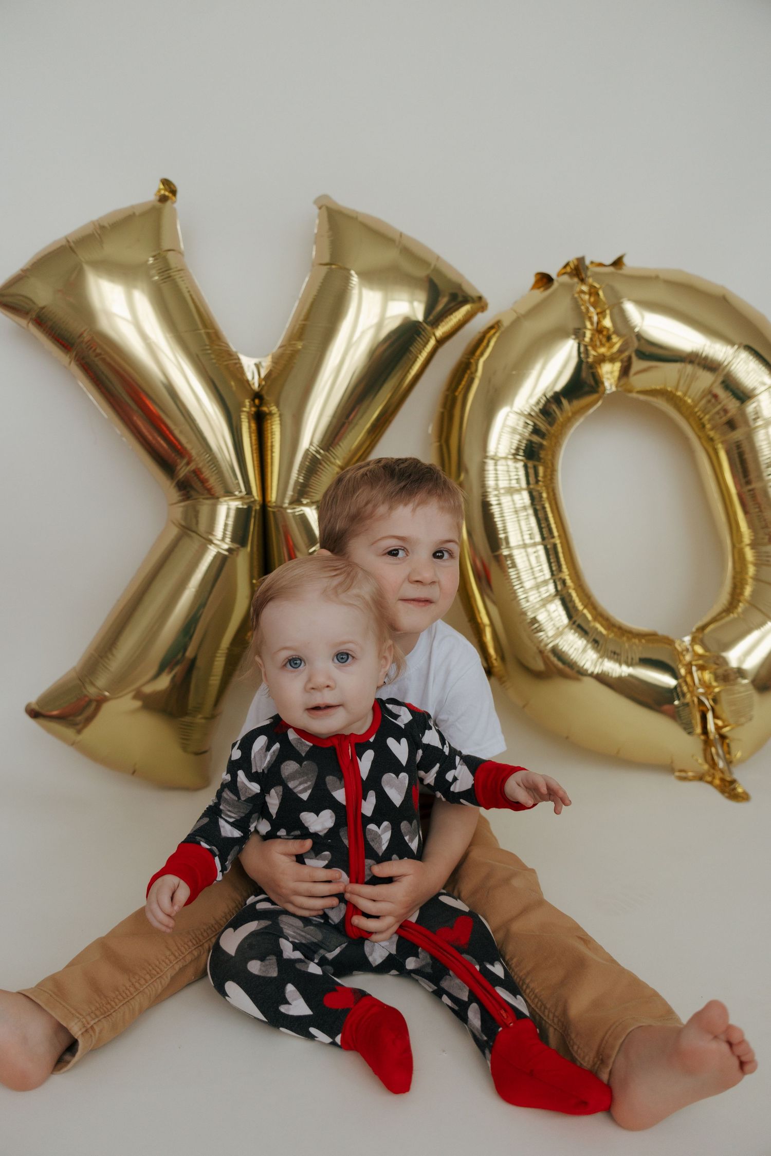 Two young children sit together in front of large gold XO balloon letters against a white backdrop.
