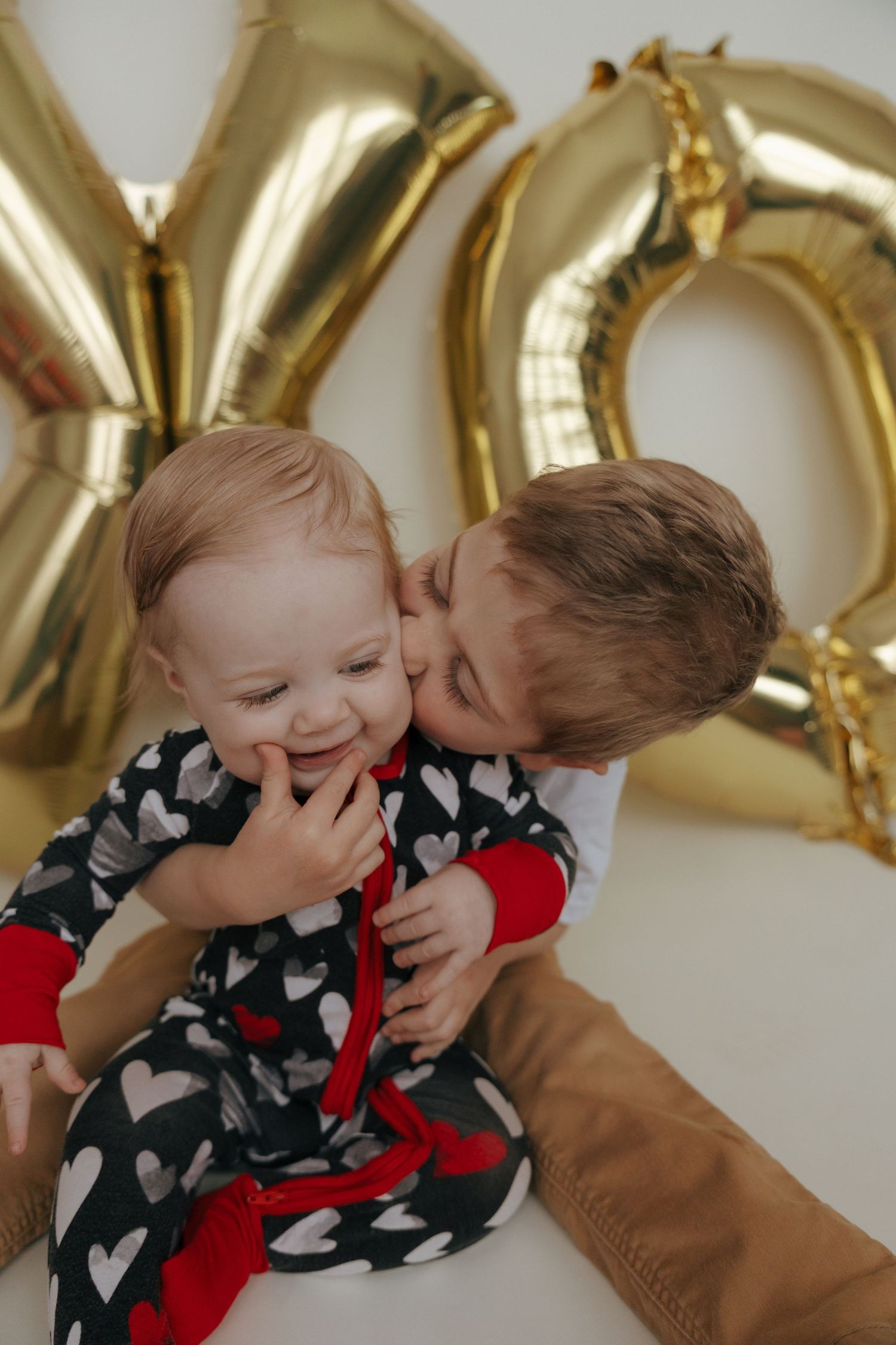 Sweet Valentine's moment with gold XO balloons and heart-patterned pajamas against white background.