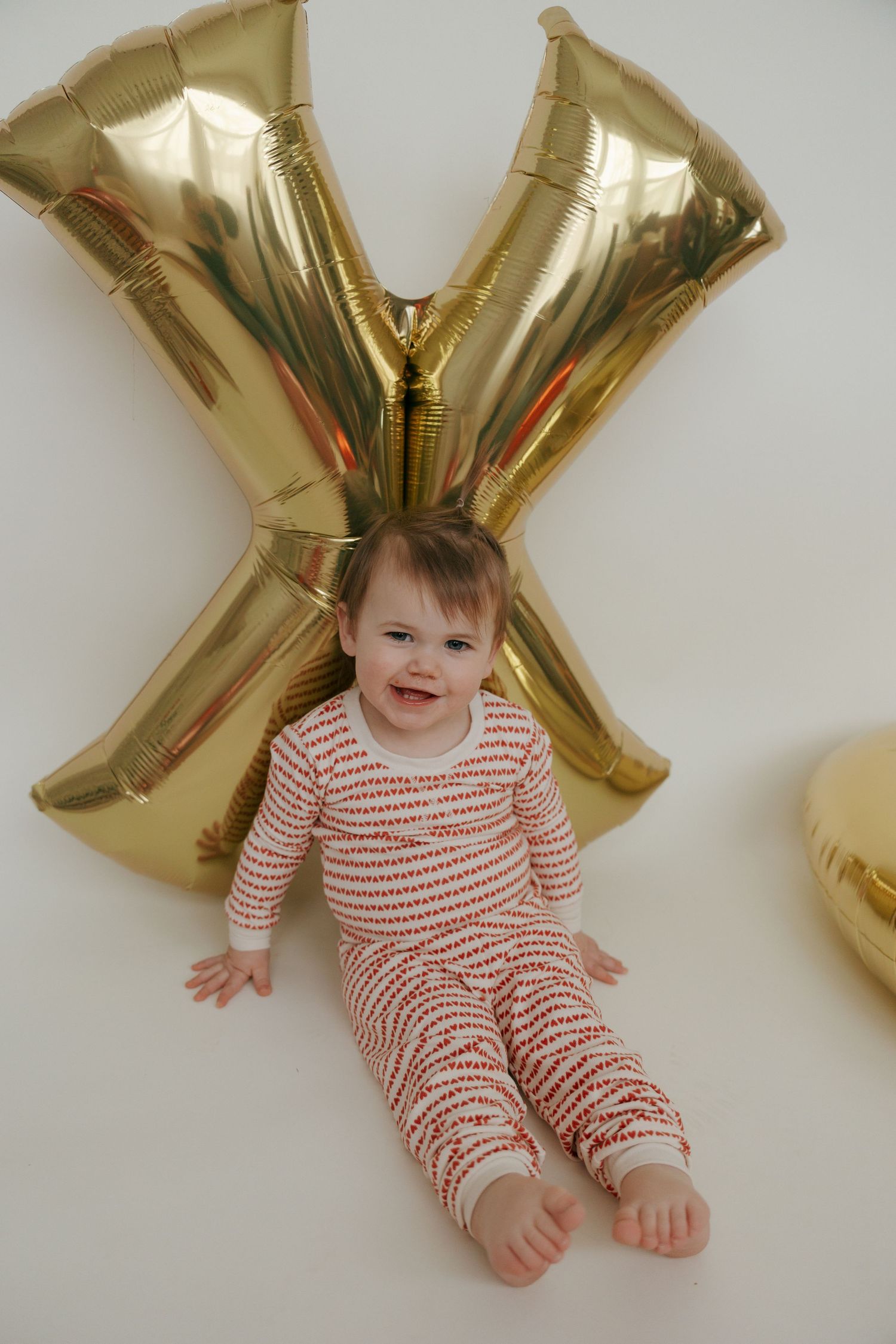 Baby wearing red and white striped pajamas lying on floor with gold letter X balloons in background.