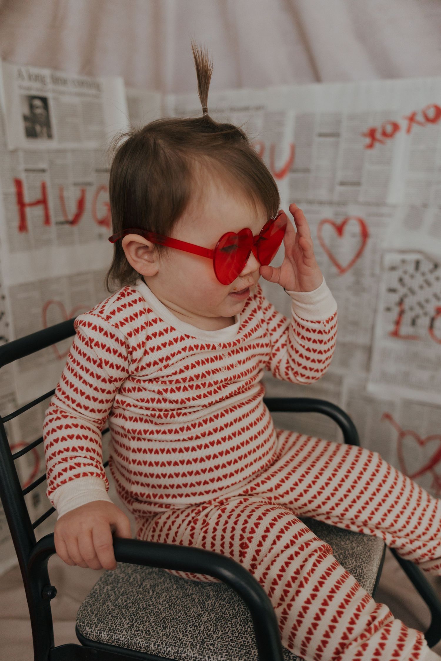 Small child wearing red heart sunglasses and striped outfit sits against newspaper-covered wall with red hearts.