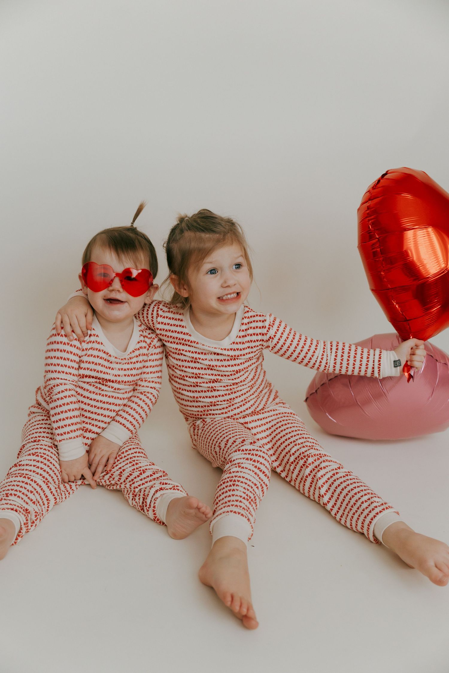 Children in matching red and white striped outfits play with heart balloons on white background.