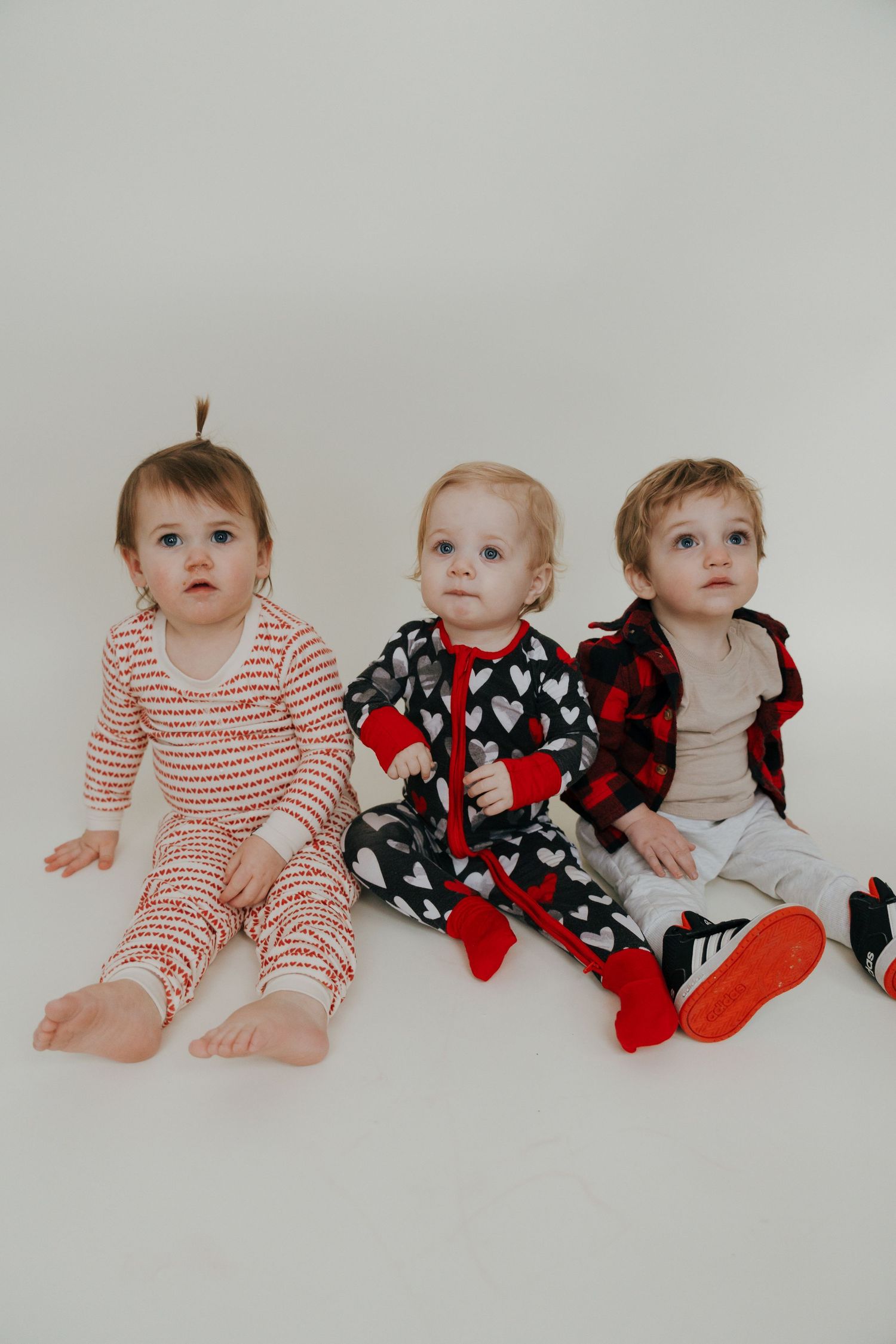 Three small children pose together wearing Valentine's themed outfits with hearts and stripes.